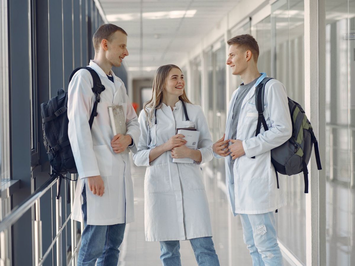 Three medical students in white coats talking in a sunlit hallway